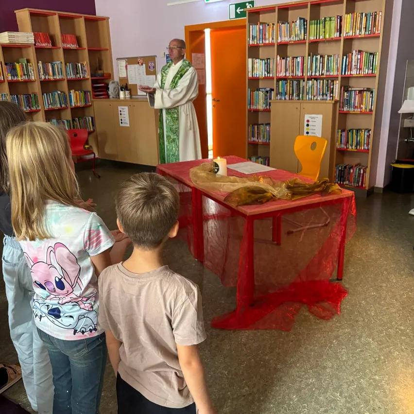 Ein Priester spricht zu einer Gruppe von Kindern in einer Bibliothek. Er steht vor einem Tisch, der mit rotem Stoff bedeckt ist.