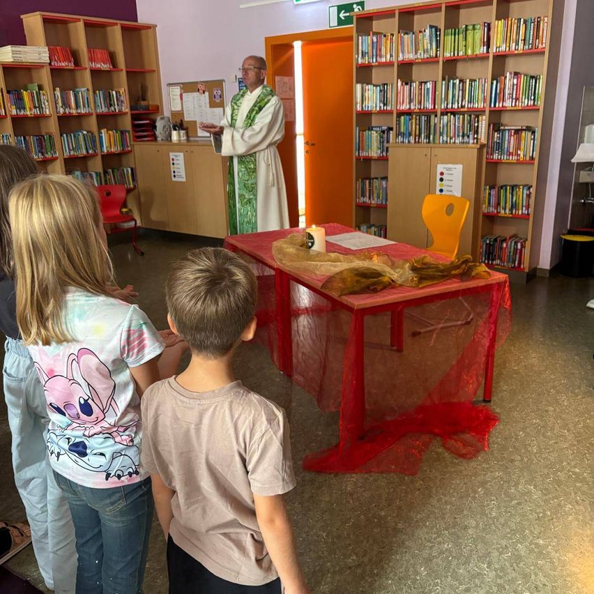 Ein Priester spricht zu einer Gruppe von Kindern in einer Bibliothek. Er steht vor einem Tisch, der mit rotem Stoff bedeckt ist.
