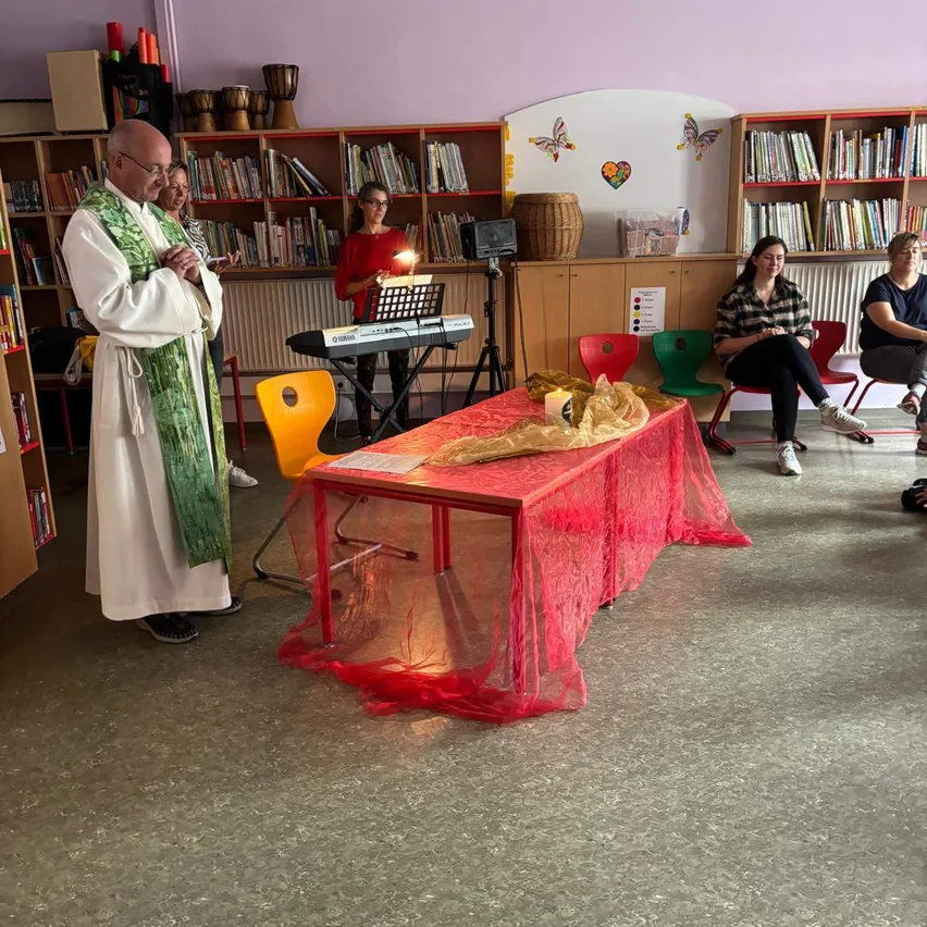Ein Priester betet in einem Klassenzimmer mit einem roten Tisch, der mit Spitze bedeckt ist. Hinter ihm spielt eine Frau Klavier. Andere Leute sitzen auf Stühlen im Raum, der mit Bücherregalen gefüllt ist.