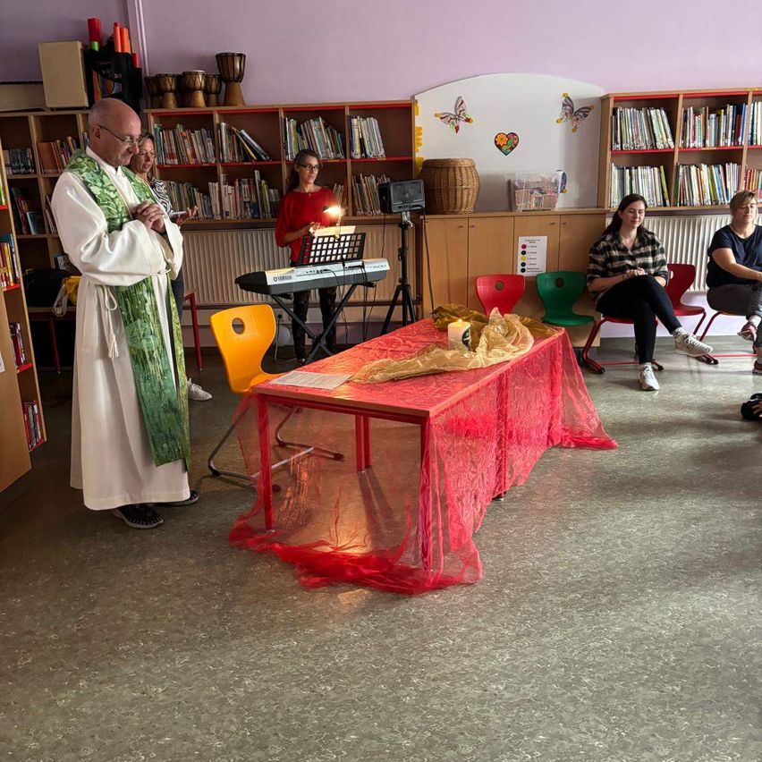 Ein Priester betet in einem Klassenzimmer mit einem roten Tisch, der mit Spitze bedeckt ist. Hinter ihm spielt eine Frau Klavier. Andere Leute sitzen auf Stühlen im Raum, der mit Bücherregalen gefüllt ist.