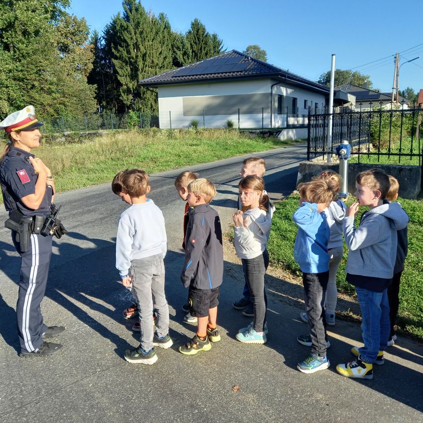 Eine Polizistin spricht mit einer Gruppe von Kindern draußen. Sie stehen auf der Straße, mit einem Haus, Bäumen und einem Zaun im Hintergrund.