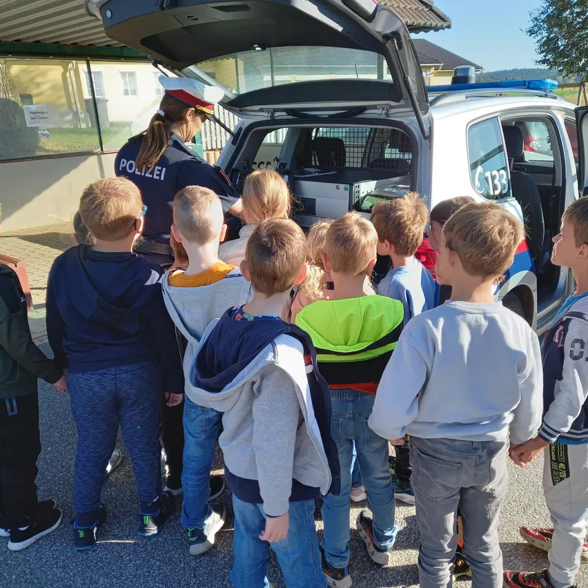 Eine Polizistin steht vor einem Polizeiauto mit offenem Kofferraum, umgeben von Kindern, die aufmerksam zuhören.