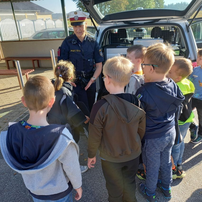 Eine Polizeibeamtin spricht mit einer Gruppe junger Kinder vor einem Polizeifahrzeug mit offenem Kofferraum. Die Kinder tragen Freizeitkleidung.