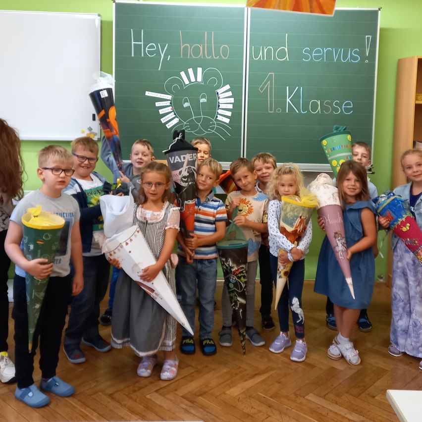 Eine Gruppe junger Kinder, in verschiedenen Outfits gekleidet, posiert für ein Foto vor einer grünen Tafel mit deutscher Schrift. Sie halten Papierkegel mit Zeichnungen und scheinen sich in einem Klassenzimmer zu befinden.