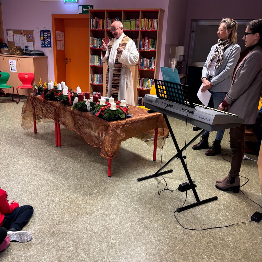 Ein Priester spricht vor einem Tisch mit Kerzen, während zwei Frauen neben einem Klavier stehen. Dahinter befindet sich ein Bücherregal und eine offene Tür.