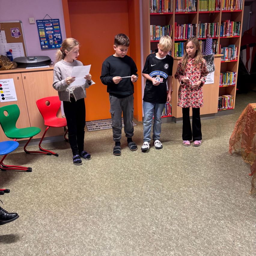 Vier Kinder stehen in einem Klassenzimmer, jedes hält ein Papier in der Hand. Ein mit Büchern gefülltes Bücherregal steht hinter ihnen.
