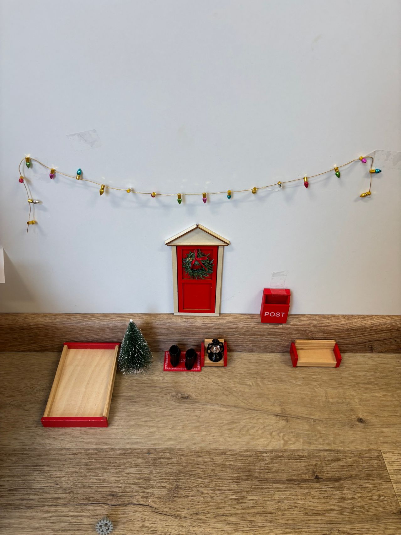Ein weihnachtlich gestalteter Spielbereich mit einer kleinen roten Tür, einem Weihnachtsbaum und einem Miniatur-Briefkasten mit der Aufschrift 'POST'. Der Boden ist aus Holz, und eine Lichterkette hängt an der Wand.