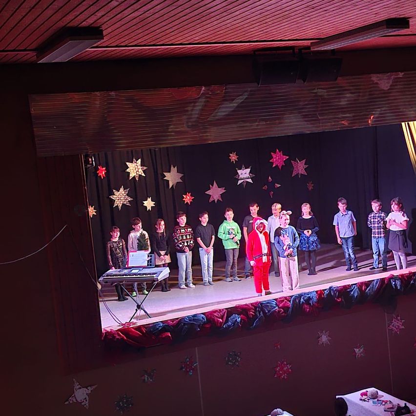 Eine Gruppe von Kindern und Erwachsenen tritt auf der Bühne auf, gekleidet in festliche Kleidung. Die Bühne ist mit Sternen und Weihnachtsdekoration geschmückt. Ein Kind spielt Klavier, während andere um ihn herum stehen.