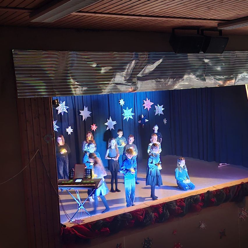 Kinder in Kostümen führen auf einer Bühne mit blauem Hintergrund und Sternen auf, halten Kerzen. Links steht ein Keyboard, und die Bühne ist mit roten und weißen Ornamenten dekoriert.