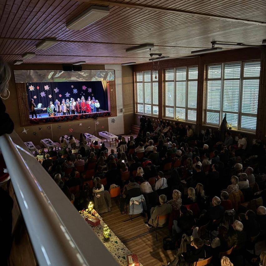 Ein überfüllter Saal mit einer Bühne an einem Ende, auf der Kinder auftreten. Viele Menschen sitzen in Stühlen und einige stehen. Tische mit Dekorationen stehen davor.