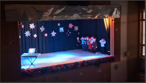 Eine dekorierte Bühne mit einer Gruppe von Menschen in Weihnachtsmützen und Winterkleidung, mit einem Klavier links und Sternen im Hintergrund.