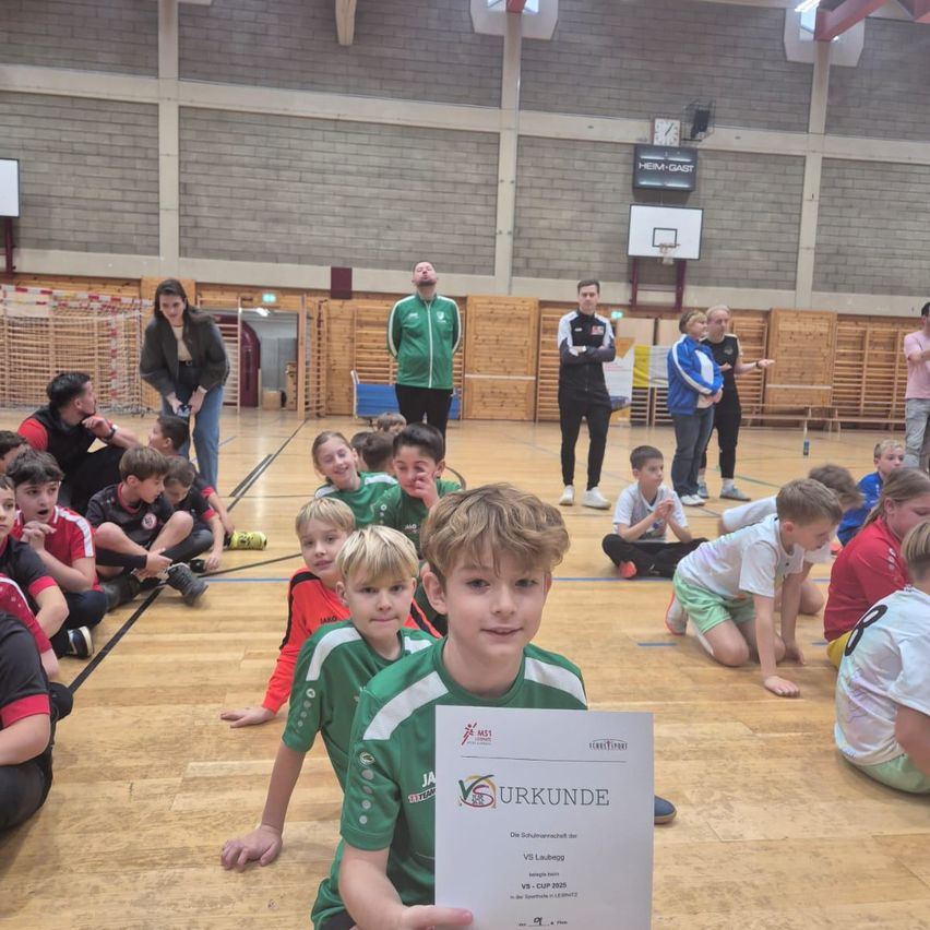 In einer Sporthalle sitzen Kinder in Sportbekleidung auf dem Boden. Ein Junge hält ein Zertifikat mit dem Wort 'URKUNDE'. Hinter ihnen stehen einige Leute, und eine Uhr ist an der Wand montiert.