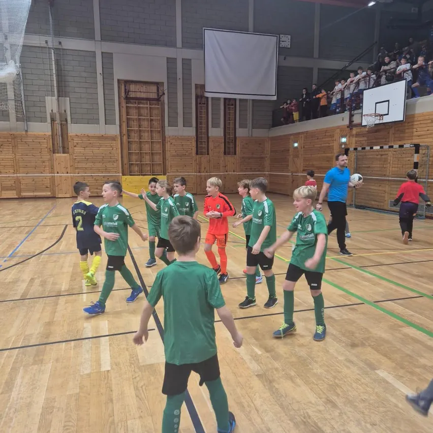 Eine Gruppe junger Jungen in grünen Trikots spielt Fußball auf einem Hallenplatz, ein Mann hält einen Ball. Zuschauer beobachten vom Balkon.