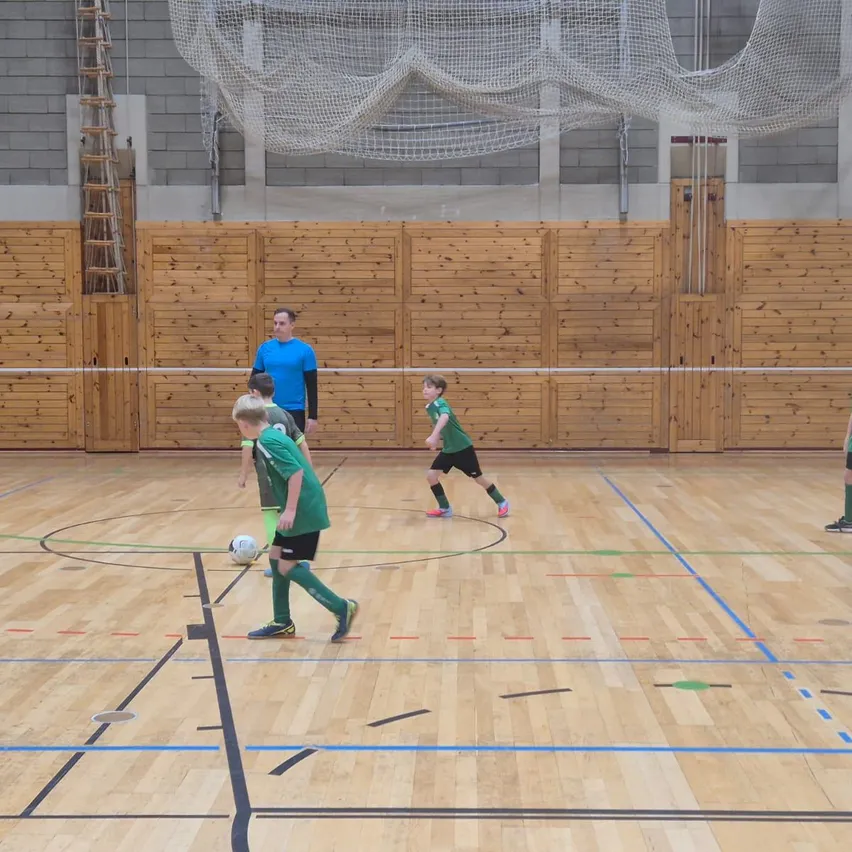 In einer Halle spielen mehrere Jungen in grünen Trikots Fußball, einer läuft auf den Ball zu. Ein Mann in einem blauen Shirt beobachtet sie. Holzwände und ein Netz sind im Hintergrund.