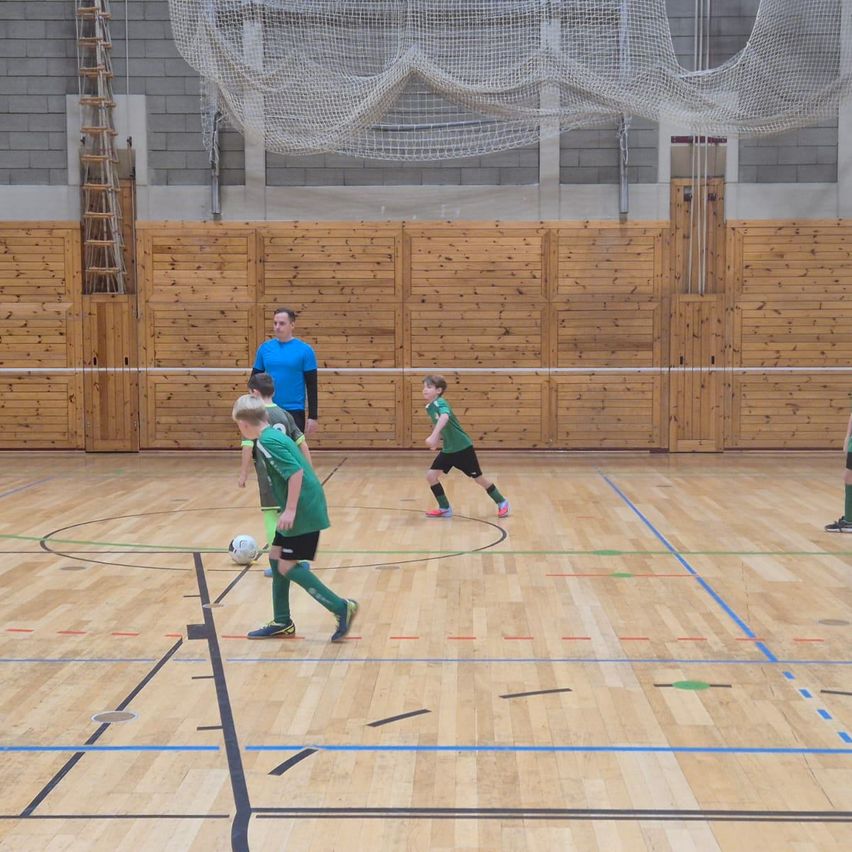 In einer Halle spielen mehrere Jungen in grünen Trikots Fußball, einer läuft auf den Ball zu. Ein Mann in einem blauen Shirt beobachtet sie. Holzwände und ein Netz sind im Hintergrund.