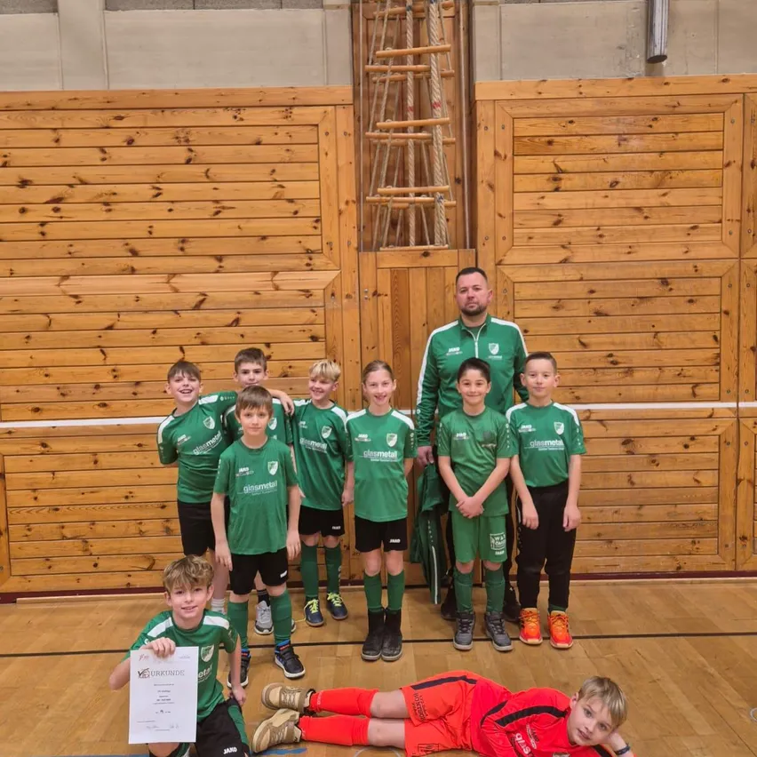 Eine Gruppe junger Fußballspieler in grünen Trikots posiert mit ihrem Trainer. Sie stehen und sitzen auf einem Holzboden. Ein Junge hält ein Zertifikat.