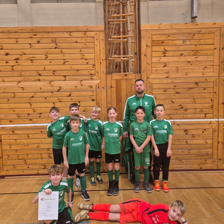 Eine Gruppe junger Fußballspieler in grünen Trikots posiert mit ihrem Trainer. Sie stehen und sitzen auf einem Holzboden. Ein Junge hält ein Zertifikat.