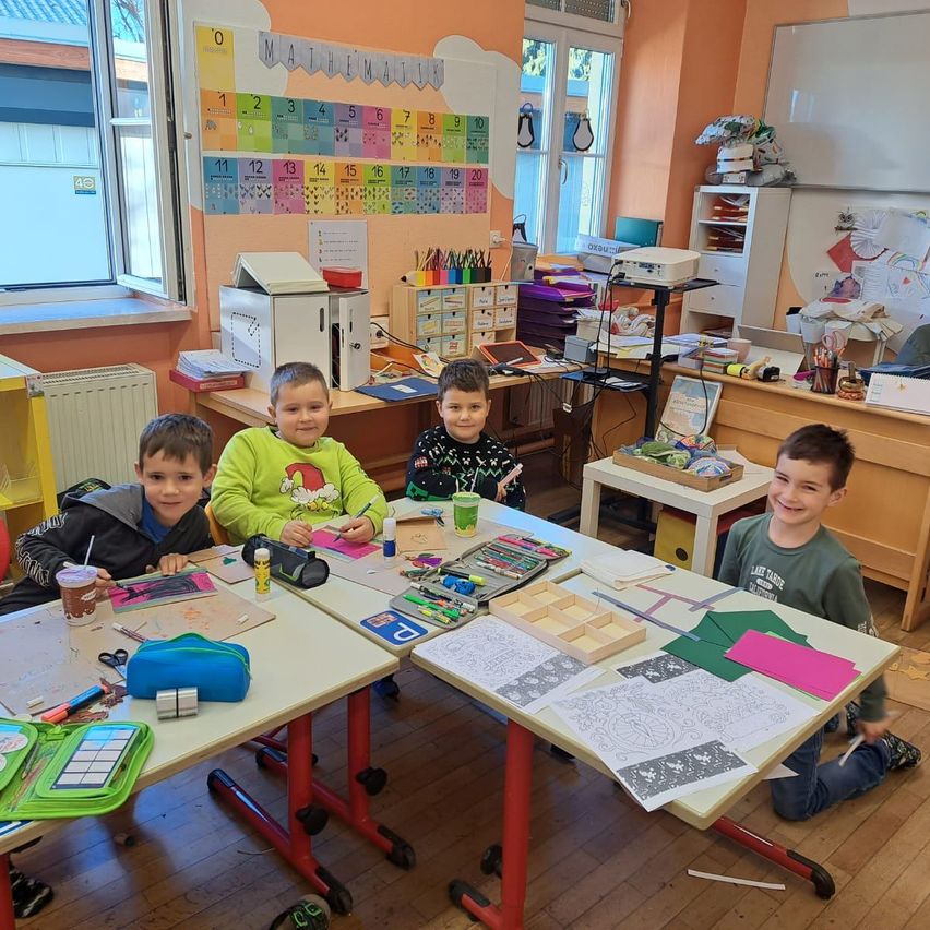 Vier Jungen sitzen an einem Tisch in einem Klassenzimmer mit verschiedenen Zeichenutensilien und Papieren darauf. Sie lächeln, und es gibt einen Mathematikplan an der Wand hinter ihnen.