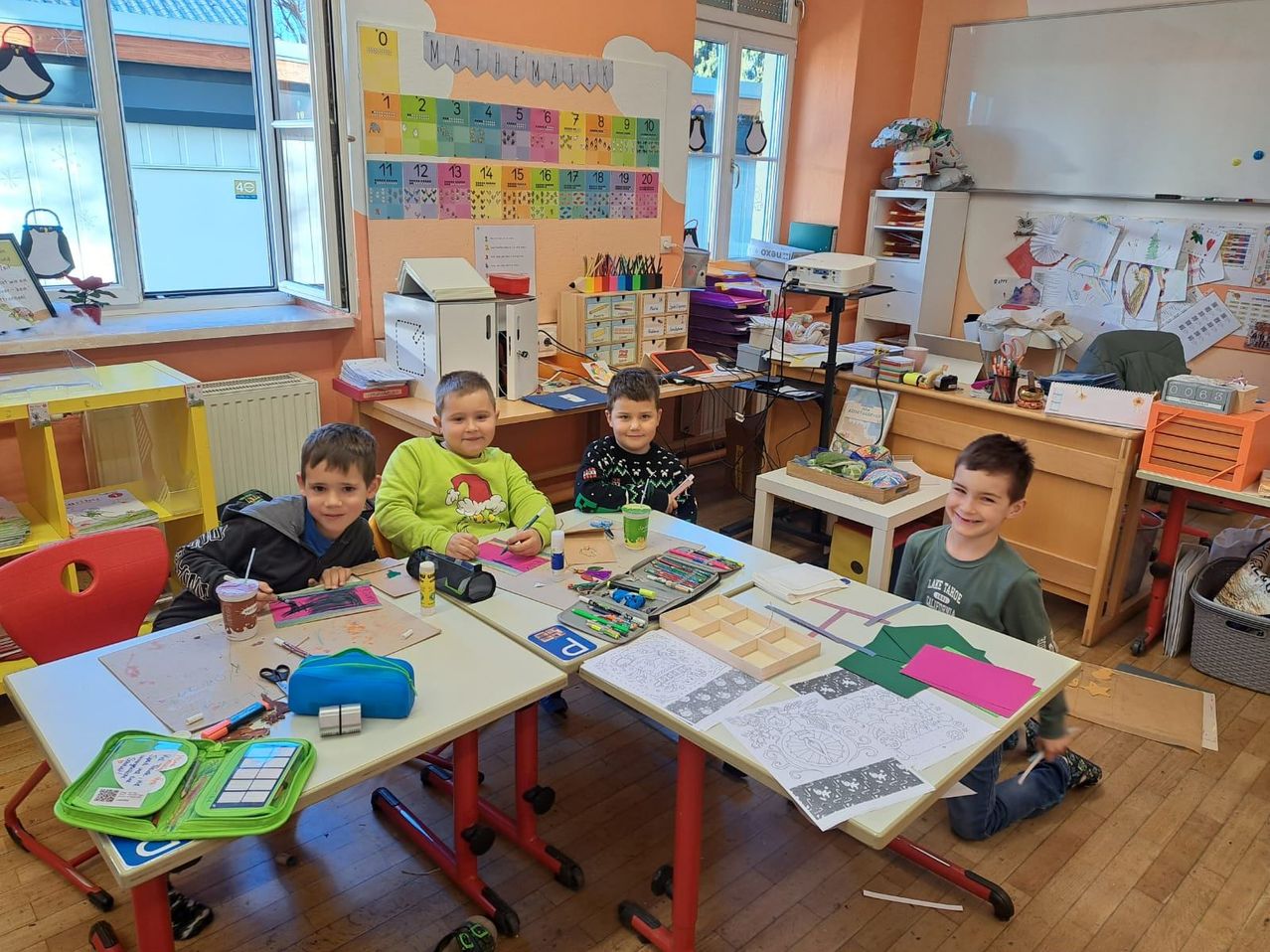 Vier Jungen sitzen an einem Tisch in einem Klassenzimmer mit verschiedenen Zeichenutensilien und Papieren darauf. Sie lächeln, und es gibt einen Mathematikplan an der Wand hinter ihnen.