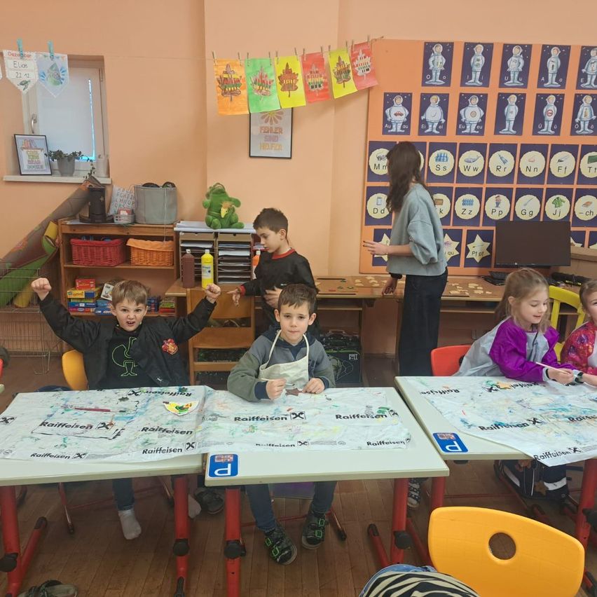 Mehrere Kinder sitzen an Tischen in einem Klassenzimmer. Eine Lehrerin steht hinter ihnen. Die Kinder sind mit Zeichen- oder Malaktivitäten beschäftigt. Das Klassenzimmer ist mit verschiedenen Postern und Dekorationen geschmückt.