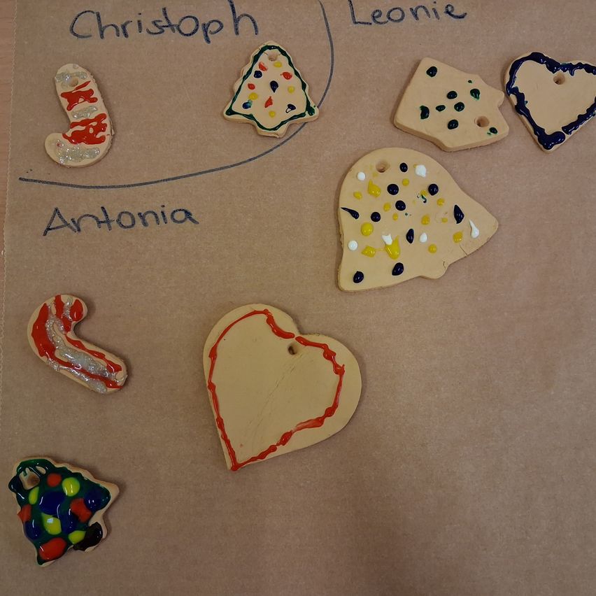 Eine Sammlung von Keramik-Anhängern wird auf einem braunen Papierhintergrund ausgestellt. Sie umfassen ein Herz, einen Weihnachtsbaum, eine Zuckerstange und andere festliche Formen. Die Namen Christoph, Leonie und Antonia sind auf dem Papier geschrieben.