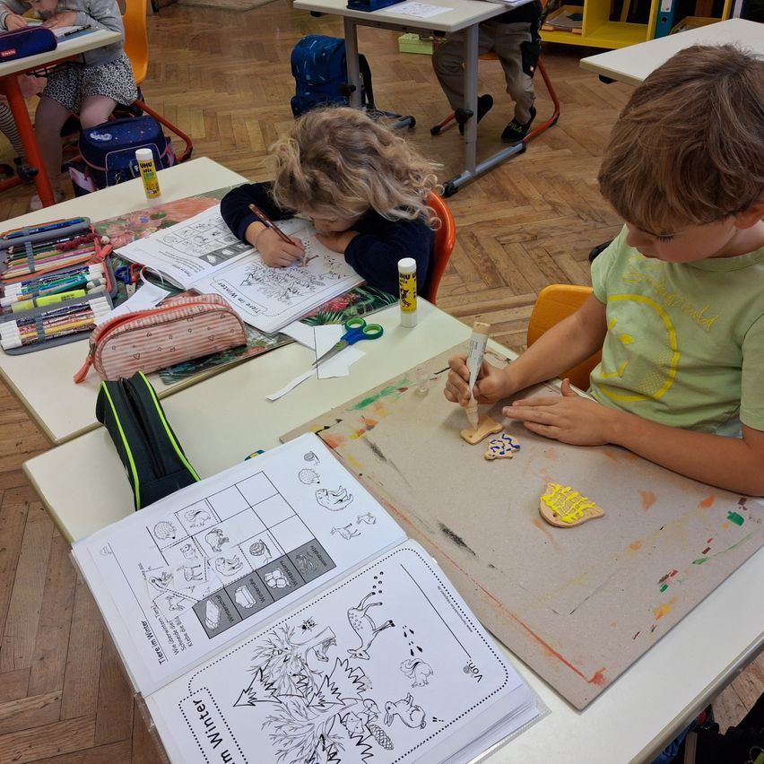 Ein Klassenzimmer mit Holzböden, Schüler zeichnen und malen, einer färbt ein Buch ein, der andere verziert Kekse, mit verschiedenen Malutensilien auf ihren Tischen.