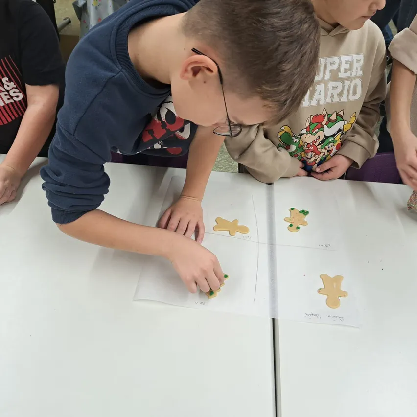 Ein Junge mit Brille beugt sich über einen Tisch und arbeitet an einer Lebkuchenmann-Aktivität. Neben ihm steht ein weiteres Kind in einem Super Mario Sweatshirt. Sie sind in einem Raum mit anderen Kindern.