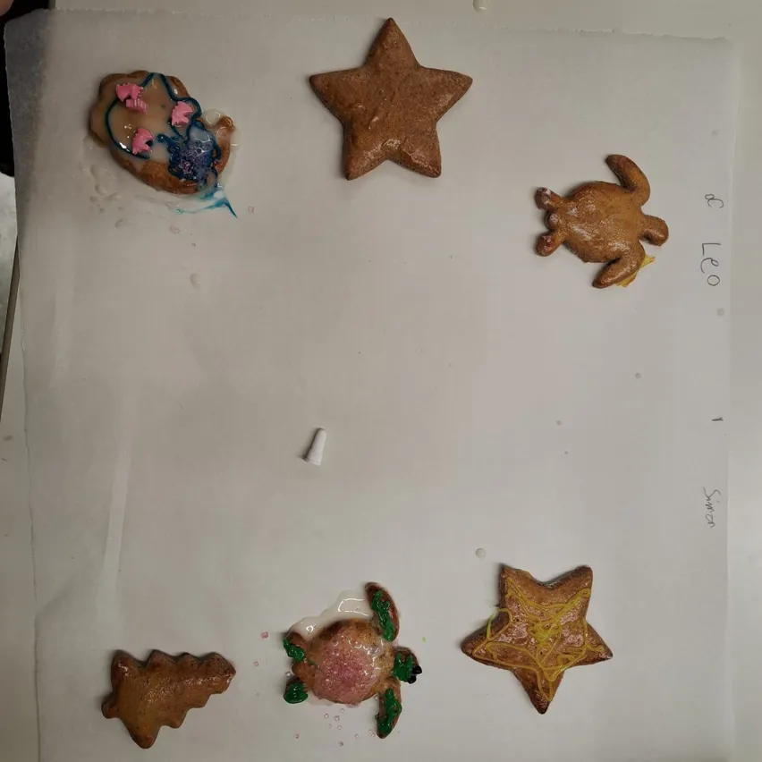 Mehrere Lebkuchen-Plätzchen in Form von einem Wal, einem Stern, einer Schildkröte, einem Weihnachtsbaum und einem Stern mit gelber Glasur liegen auf einer weißen Oberfläche. Einige haben blaue und grüne Glasur.