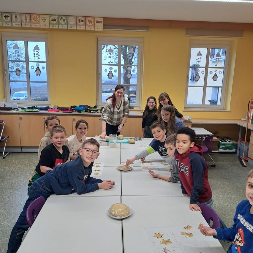 Ein Klassenzimmer voller Kinder an einem Tisch, einige stehen, andere sitzen, mit einer Frau, die hinter ihnen steht. Der Raum hat gelbe Wände, Fenster mit Pinguindekorationen und ein Baum ist draußen sichtbar.