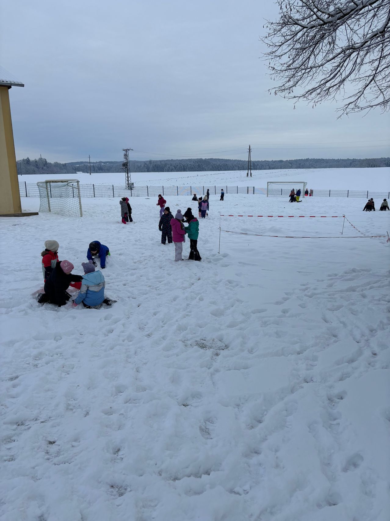 Kinder spielen im Schnee, einige stehen herum und andere sitzen. Links befinden sich zwei Fußballtore. Schnee bedeckt den Boden, und ein Zaun umgibt das Gebiet. In der Ferne gibt es Bäume und einen klaren Himmel.