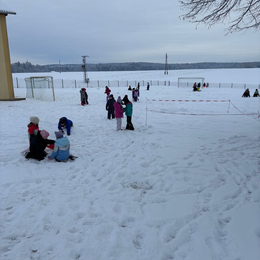 Kinder spielen im Schnee, einige stehen herum und andere sitzen. Links befinden sich zwei Fußballtore. Schnee bedeckt den Boden, und ein Zaun umgibt das Gebiet. In der Ferne gibt es Bäume und einen klaren Himmel.