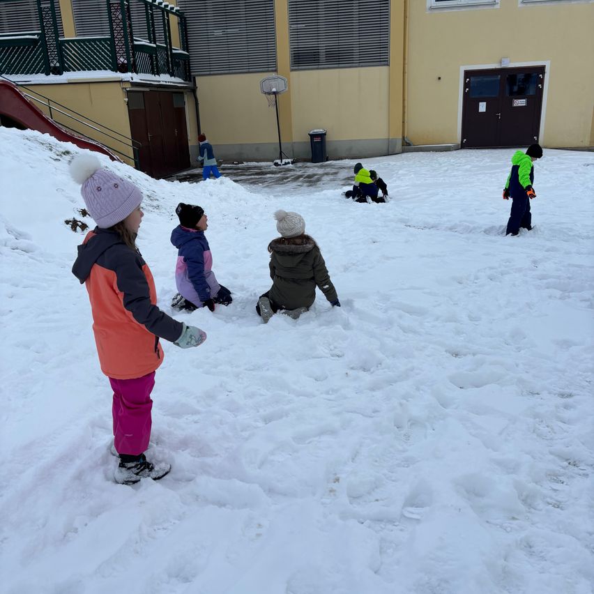 Mehrere Kinder spielen im Schnee vor einem Schulgebäude mit einem Basketballkorb. Sie tragen Winterkleidung und Handschuhe.