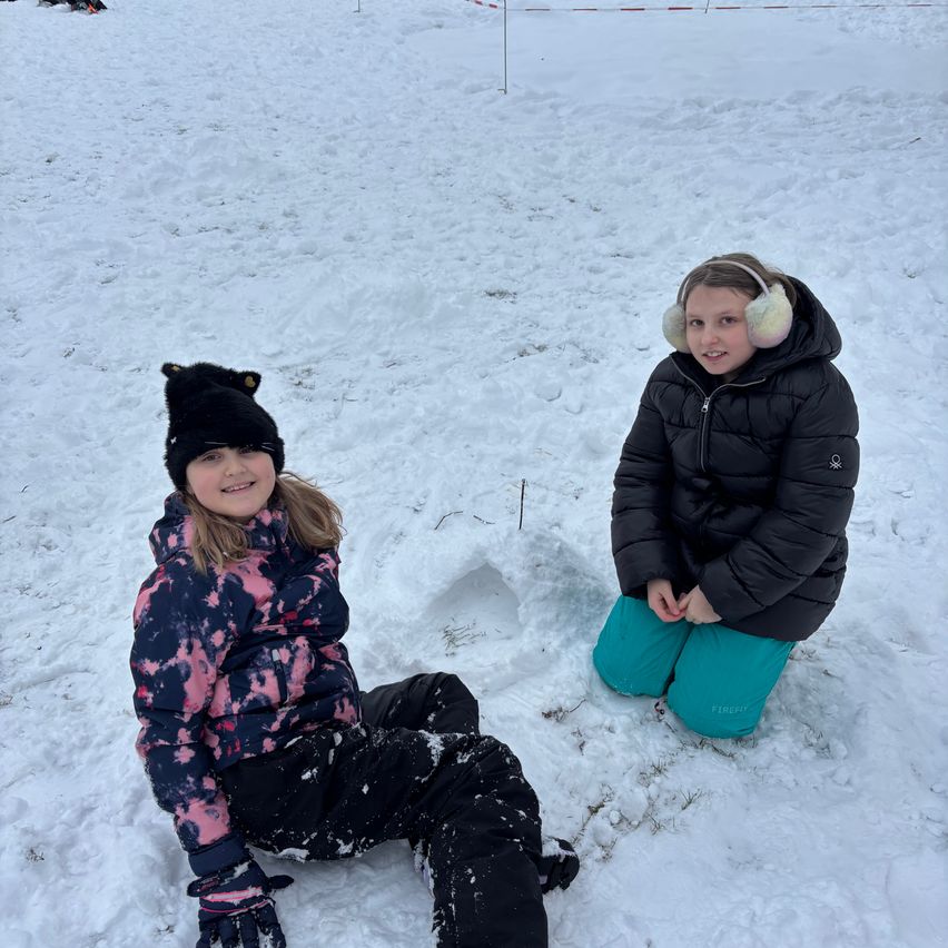Zwei Mädchen sitzen im Schnee und lächeln beide. Das Mädchen auf der linken Seite trägt eine Tie-Dye-Jacke und Handschuhe. Das Mädchen auf der rechten Seite trägt eine schwarze Jacke mit weißen Ohrenschützern. Schnee ist um sie herum verstreut.