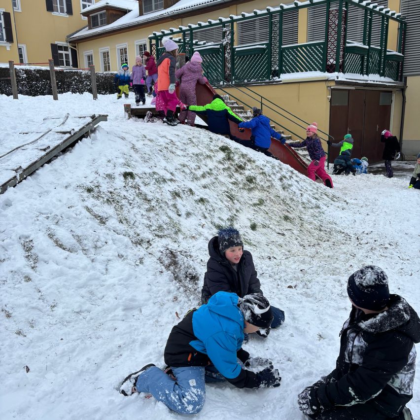 Kinder spielen im Schnee vor einem Gebäude mit einer roten Rutsche. Einige klettern die Rutsche hinauf, andere sitzen und spielen im Schnee.