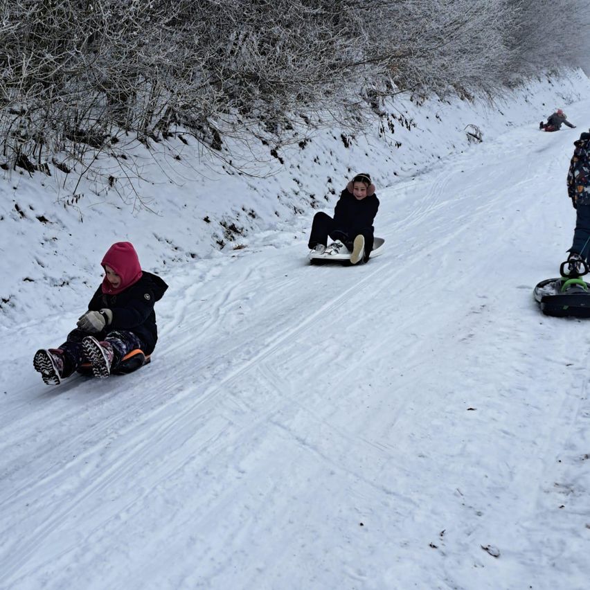 Mehrere Kinder genießen das Schlittenfahren auf einem verschneiten Hang, eines von ihnen trägt eine pinke Kapuze und eine Brille.