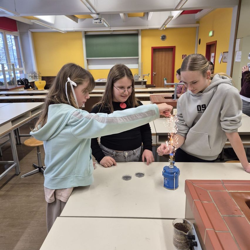 Drei Mädchen in einem Chemieunterricht führen ein wissenschaftliches Experiment durch. Ein Mädchen pustet auf einen Funkensprühenden Gegenstand, während ein anderes Mädchen einen blauen Kanister hält.