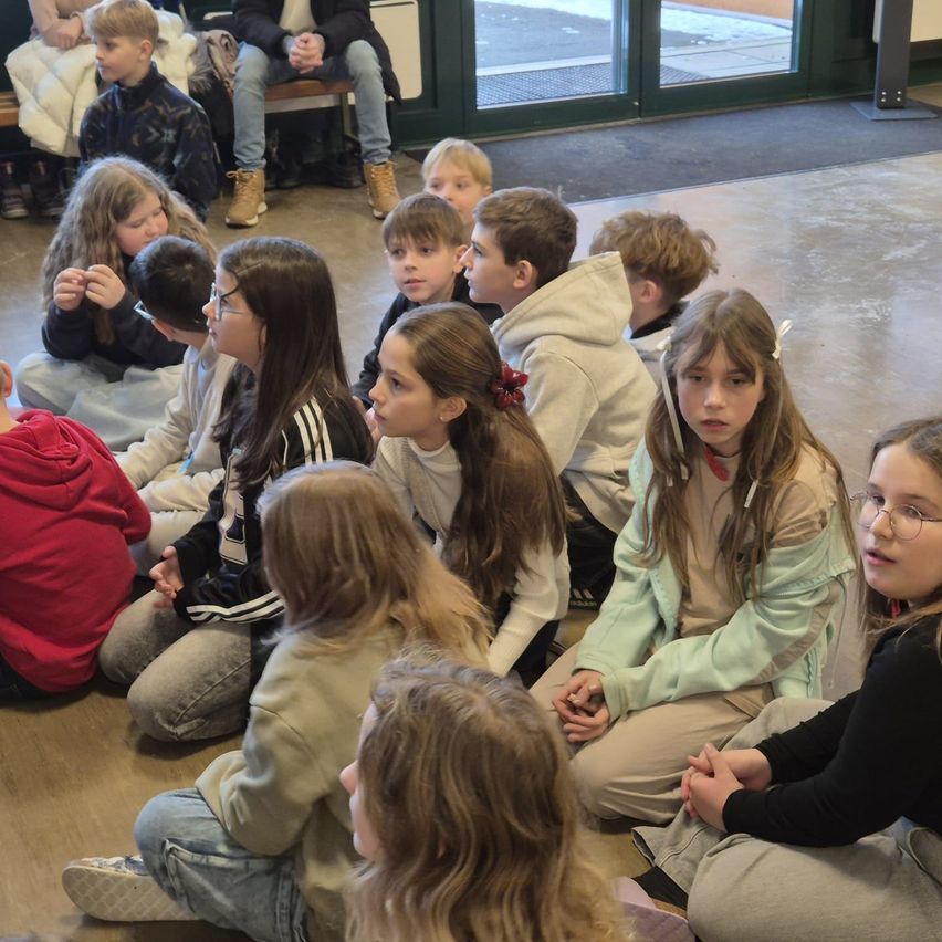 Eine Gruppe von Kindern sitzt auf dem Boden, möglicherweise in einem Klassenzimmer. Einige tragen Brillen, Jacken und Turnschuhe. Sie scheinen einer Person vor ihnen zuzuhören.