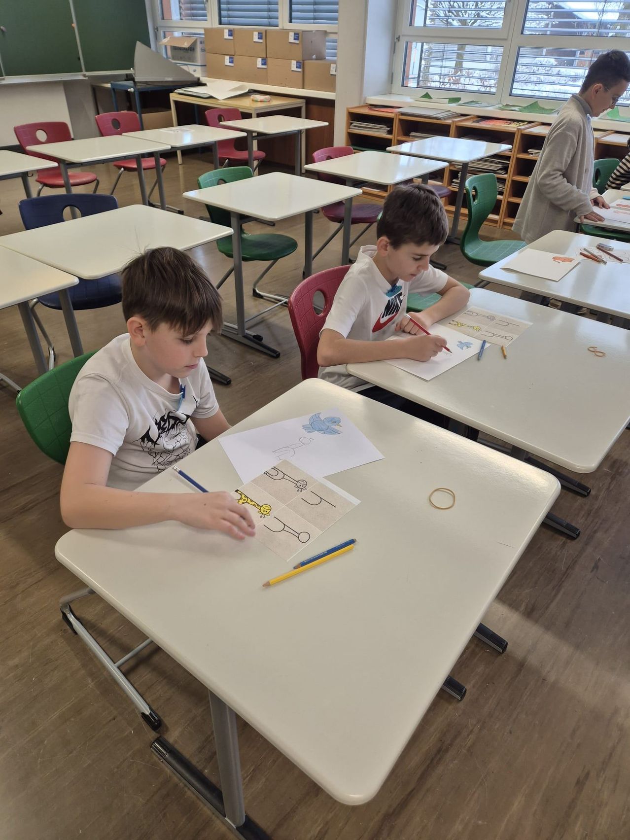 Zwei Jungen sitzen an Schreibtischen in einem Klassenzimmer und sind mit Malaktivitäten beschäftigt. Sie sind von anderen Schülern und Tischen umgeben.