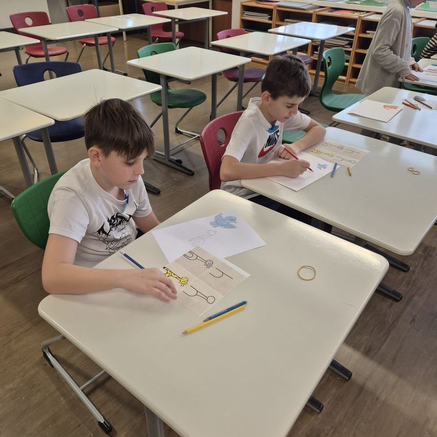 Zwei Jungen sitzen an Schreibtischen in einem Klassenzimmer und sind mit Malaktivitäten beschäftigt. Sie sind von anderen Schülern und Tischen umgeben.