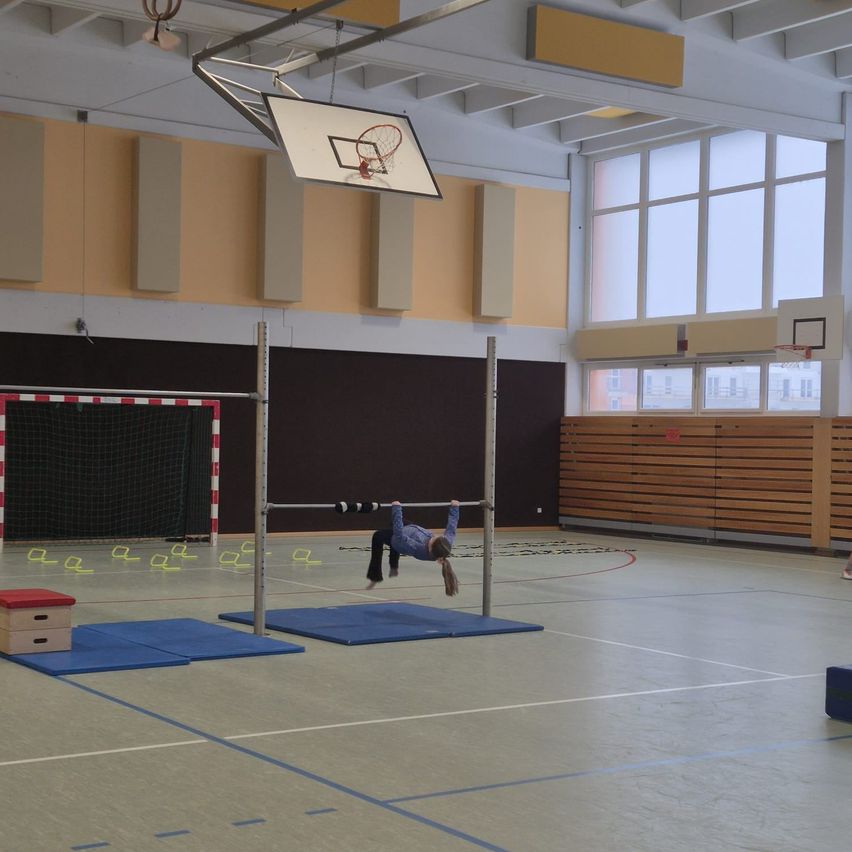 Ein junges Mädchen führt einen Handstand an horizontalen Stangen in einer Turnhalle aus, umgeben von blauen Matten und einem Basketballkorb.