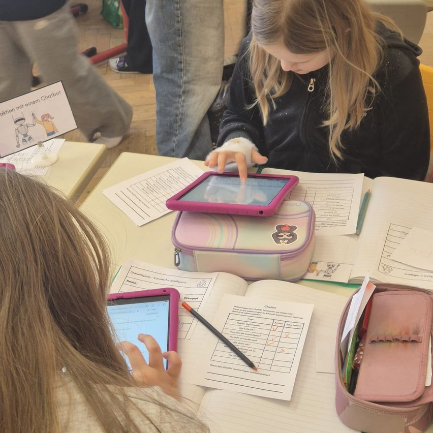 Zwei junge Mädchen arbeiten an einem Tisch mit Papieren, Stiften und einem Lunchbox an Tablets. Eine Person in Jeans steht hinter ihnen.
