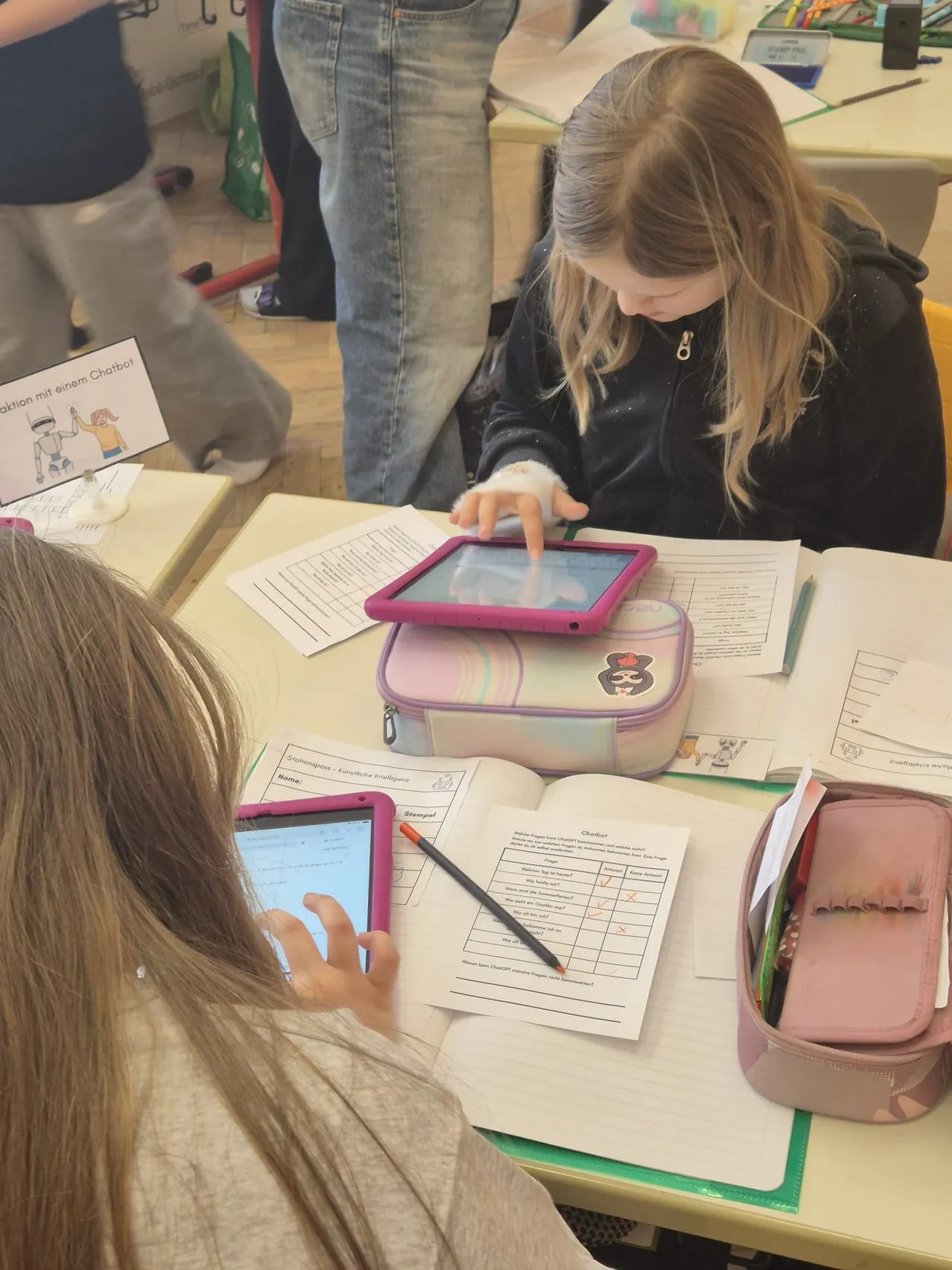 Zwei junge Mädchen arbeiten an einem Tisch mit Papieren, Stiften und einem Lunchbox an Tablets. Eine Person in Jeans steht hinter ihnen.