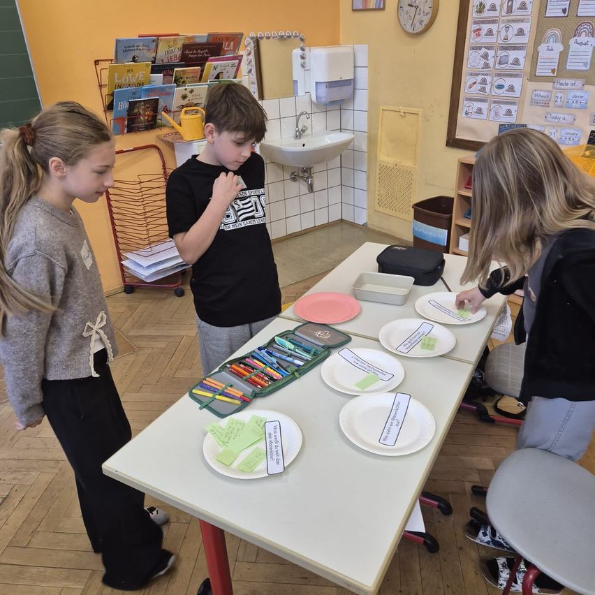 Drei Kinder stehen um einen Tisch mit Papiertellern und Markern. Ein Mädchen links trägt ein Band in den Haaren. Ein Junge in der Mitte trägt ein schwarzes T-Shirt. Ein weiteres Mädchen rechts trägt Turnschuhe.