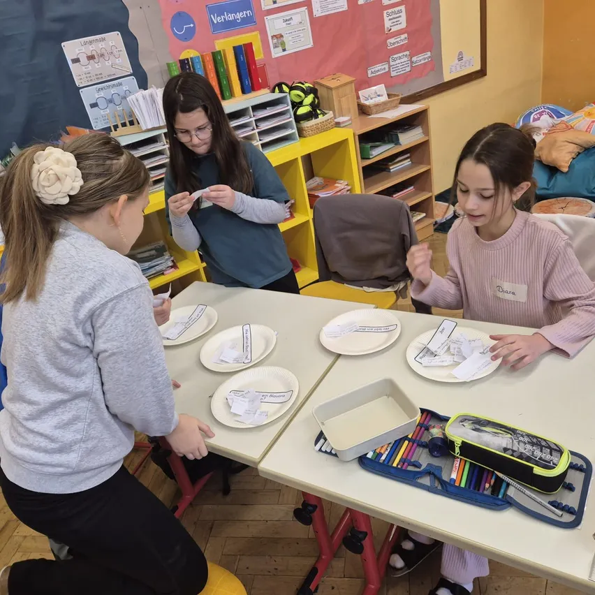 Drei junge Mädchen in einem Klassenzimmer, die an einer Aktivität mit Tellern und Papieren beteiligt sind. Das Mädchen auf der rechten Seite hat einen Namensschild mit dem Namen Diana. Dahinter steht ein Regal mit Büchern und einem Korb.
