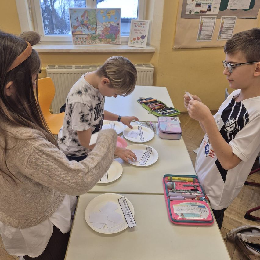 In einem Klassenzimmer beschäftigen sich drei Kinder mit einer Aktivität. Auf dem Tisch vor ihnen liegen Papierteller mit Papierausschnitten. Ein Kind trägt eine Brille.
