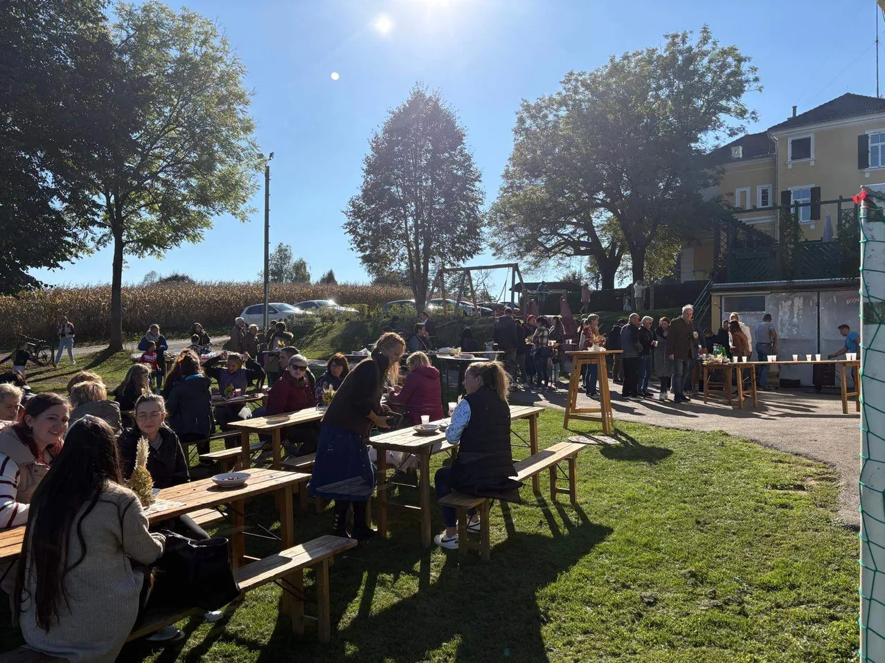 An einem sonnigen Tag versammeln sich viele Menschen in einem Außenbereich mit Holztischen und Bänken, wahrscheinlich für ein Picknick. Bäume und ein Gebäude sind im Hintergrund.