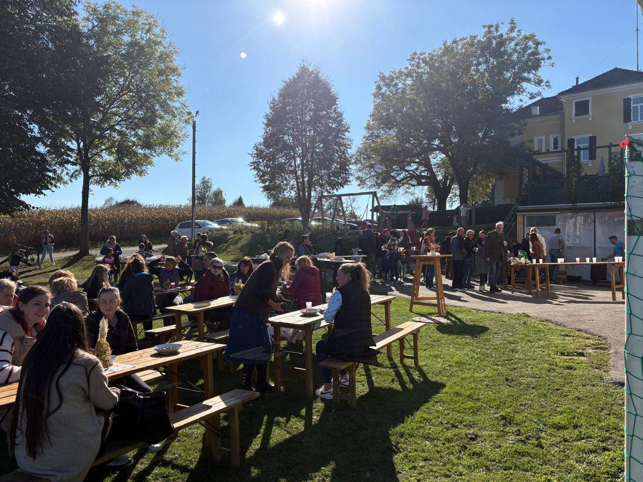 An einem sonnigen Tag versammeln sich viele Menschen in einem Außenbereich mit Holztischen und Bänken, wahrscheinlich für ein Picknick. Bäume und ein Gebäude sind im Hintergrund.