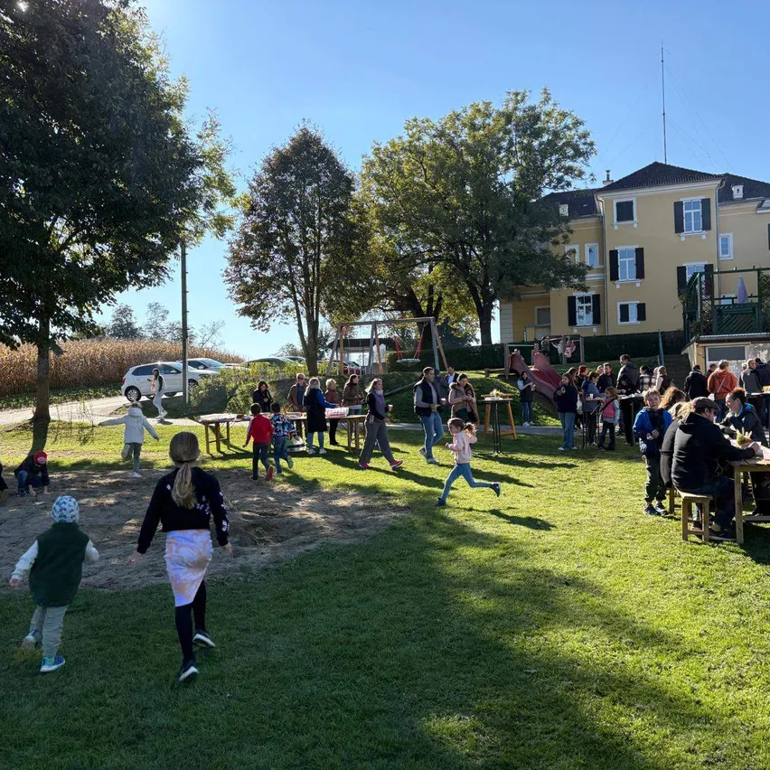 Ein sonniger Tag mit Familien und Kindern, die Outdoor-Aktivitäten genießen. Einige spielen auf dem Rasen, andere sitzen an Tischen, und ein großes Gebäude ist im Hintergrund.