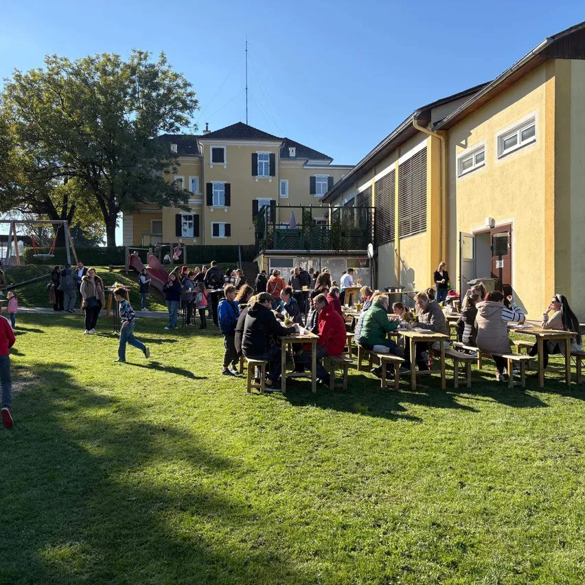Eine Gruppe von Menschen sitzt an Tischen im Freien in einem Park, während ein Kind auf dem Gras läuft. In der Nähe sind ein Baum und ein Gebäude zu sehen.