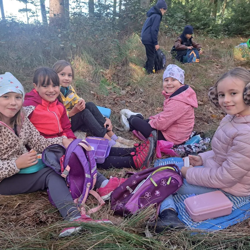 Fünf junge Mädchen sitzen auf dem Boden in einem Wald. Sie tragen Winterkleidung und haben Rucksäcke. Einige lächeln. Hinter ihnen stehen andere Leute, eine davon steht.